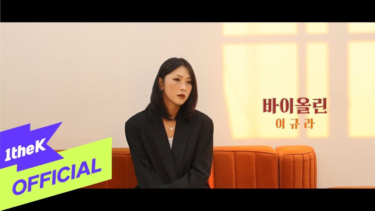 Lee KyuRa sentada em um sofá laranja, com expressão reflexiva, em um cenário minimalista e iluminado.