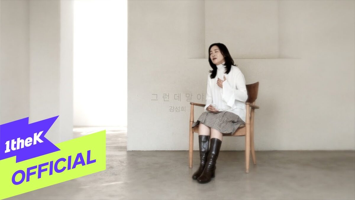 Kang Seonghee canta em uma cadeira, vestindo blusa branca e saia xadrez, em um ambiente minimalista.