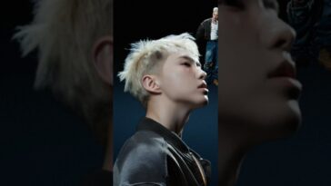 HOSHI em perfil, com cabelo loiro e expressão intensa, contra um fundo escuro e elementos de cena ao fundo.