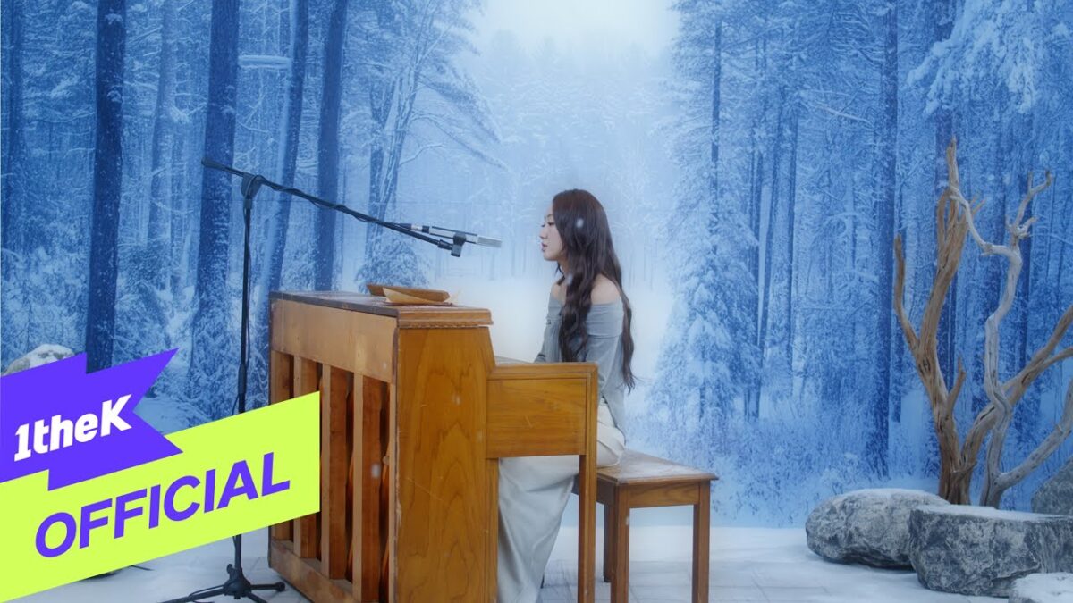 Heo Hoy Kyung canta ao piano em um cenário de inverno, com árvores cobertas de neve ao fundo.