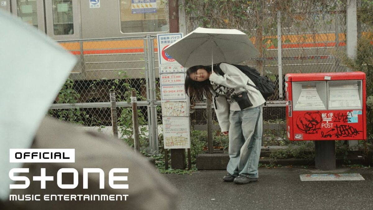 Uma jovem com um guarda-chuva se inclina levemente em uma estação de trem, ao lado de uma caixa de correio grafitada.