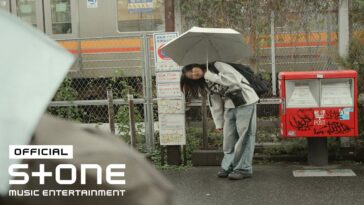 Uma jovem com um guarda-chuva se inclina levemente em uma estação de trem, ao lado de uma caixa de correio grafitada.