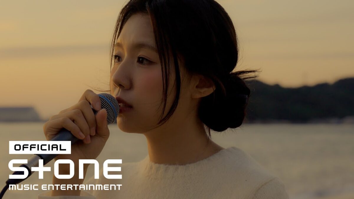 Cantora Choi Ingyeong se apresenta ao vivo com um microfone, em um cenário ao pôr do sol.
