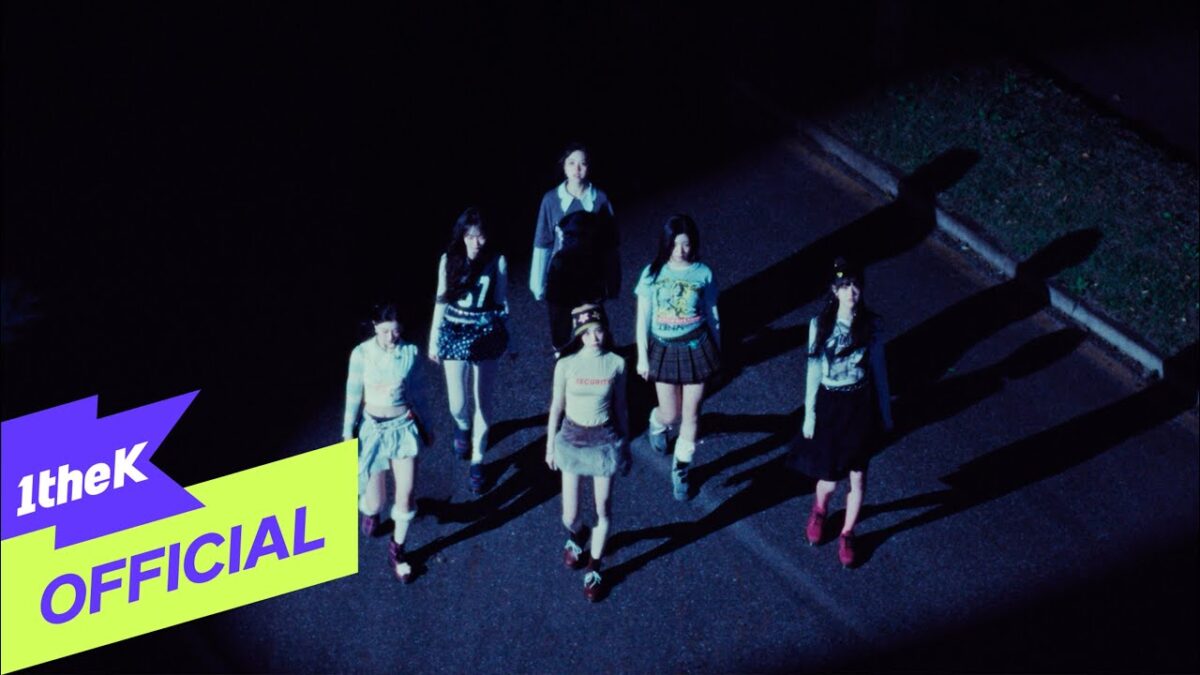 Seis jovens caminham juntas à noite, com sombras projetadas no chão, em um ambiente urbano.