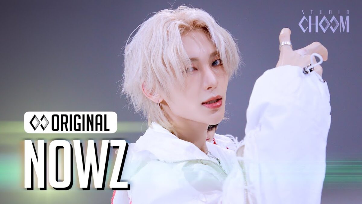 Artista com cabelo platinado e roupa branca, posando com uma expressão confiante durante apresentação.