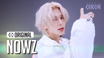 Artista com cabelo platinado e roupa branca, posando com uma expressão confiante durante apresentação.