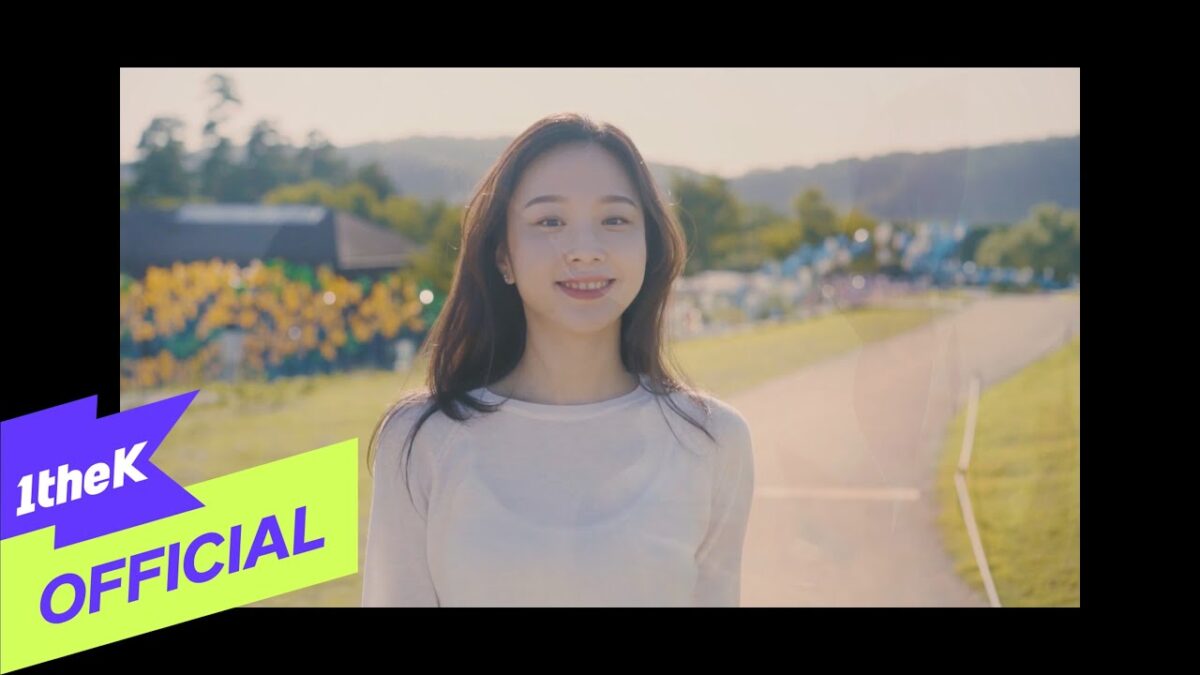 Lee Na Rae sorri enquanto caminha por um caminho em um ambiente ensolarado, com flores ao fundo.