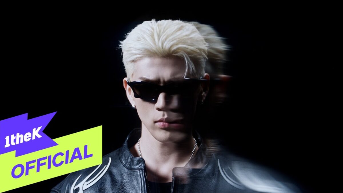 Artista com cabelo platinado e óculos escuros, posando em fundo escuro, transmite atitude e estilo no videoclipe "Ooh".
