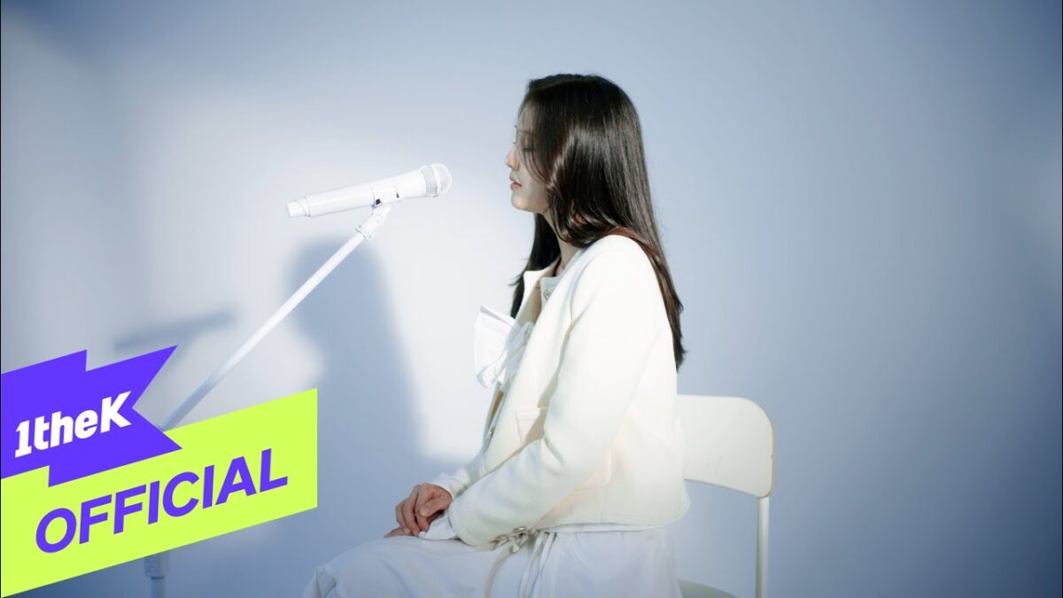 Jeong Hyo Bean sentada em uma cadeira, cantando em um microfone em um fundo minimalista.