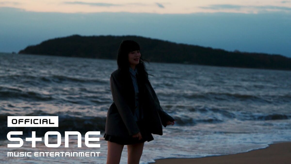 Joyce em pé na praia ao entardecer, com ondas ao fundo e um olhar sonhador.