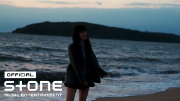 Joyce em pé na praia ao entardecer, com ondas ao fundo e um olhar sonhador.