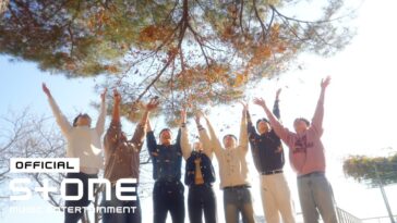 Grupo de amigos sorri e levanta as mãos para o céu, enquanto folhas caem ao seu redor em um dia ensolarado.