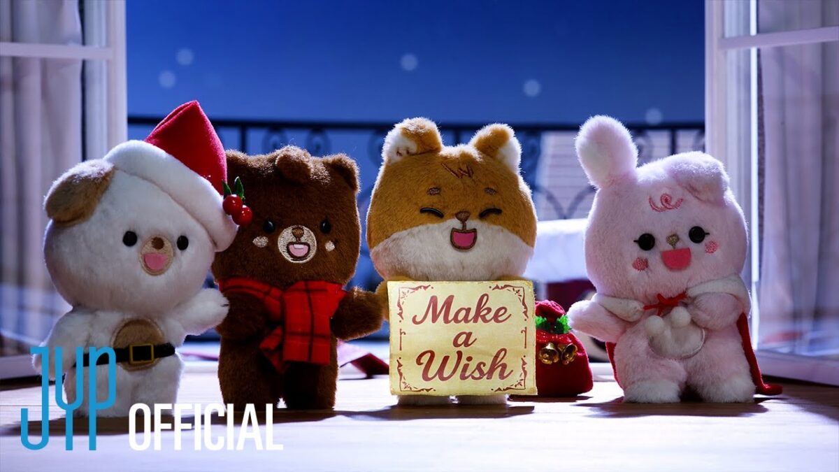 Quatro bonecos de pelúcia festivos seguram um cartaz que diz "Make a Wish" em um cenário natalino iluminado.