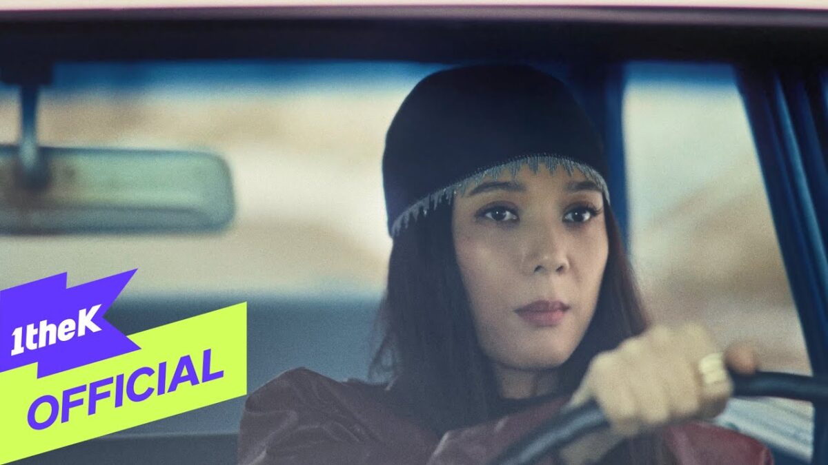 Mulher dirigindo um carro, com expressão concentrada, usando um gorro e roupas escuras.