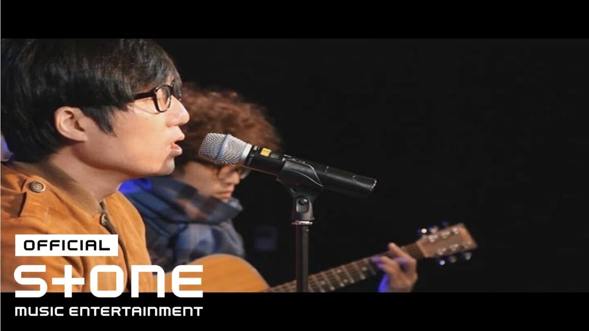 Cantor se apresenta ao microfone enquanto o guitarrista toca ao fundo, em um ambiente de palco iluminado.