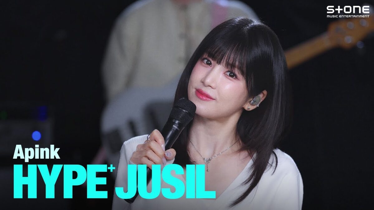 Vocalista da Apink sorri enquanto segura um microfone durante apresentação ao vivo da música "Love Me More".