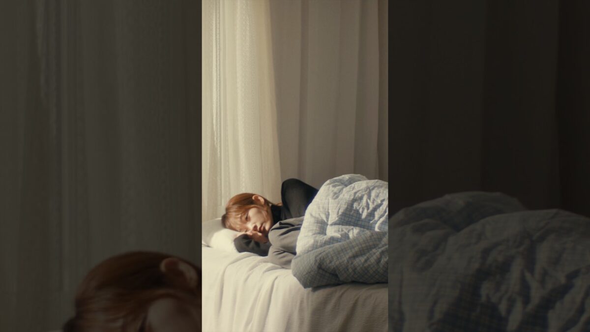 Dokyeom dorme tranquilamente em uma cama, envolto em cobertores, com luz suave entrando pela janela.