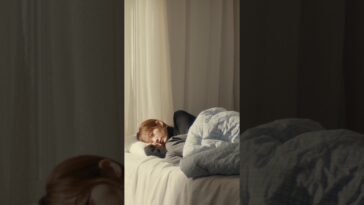 Dokyeom dorme tranquilamente em uma cama, envolto em cobertores, com luz suave entrando pela janela.