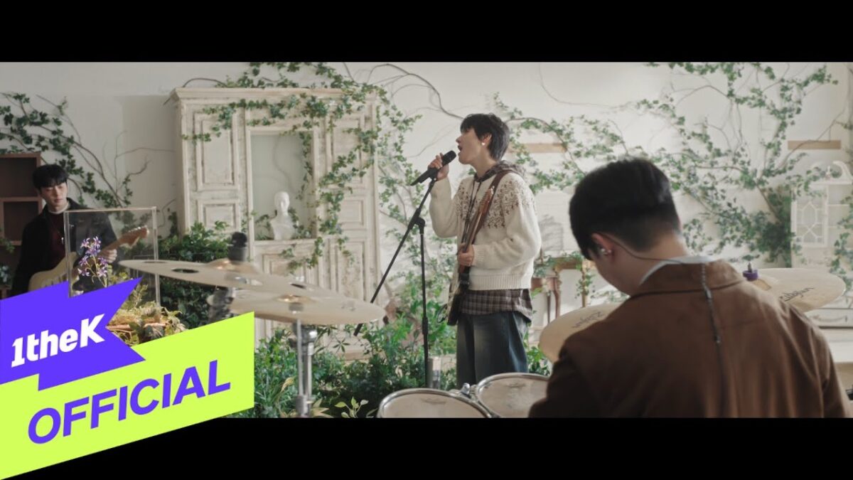 Banda ONEWE se apresenta em um ambiente decorado com plantas, enquanto um vocalista canta em um microfone.