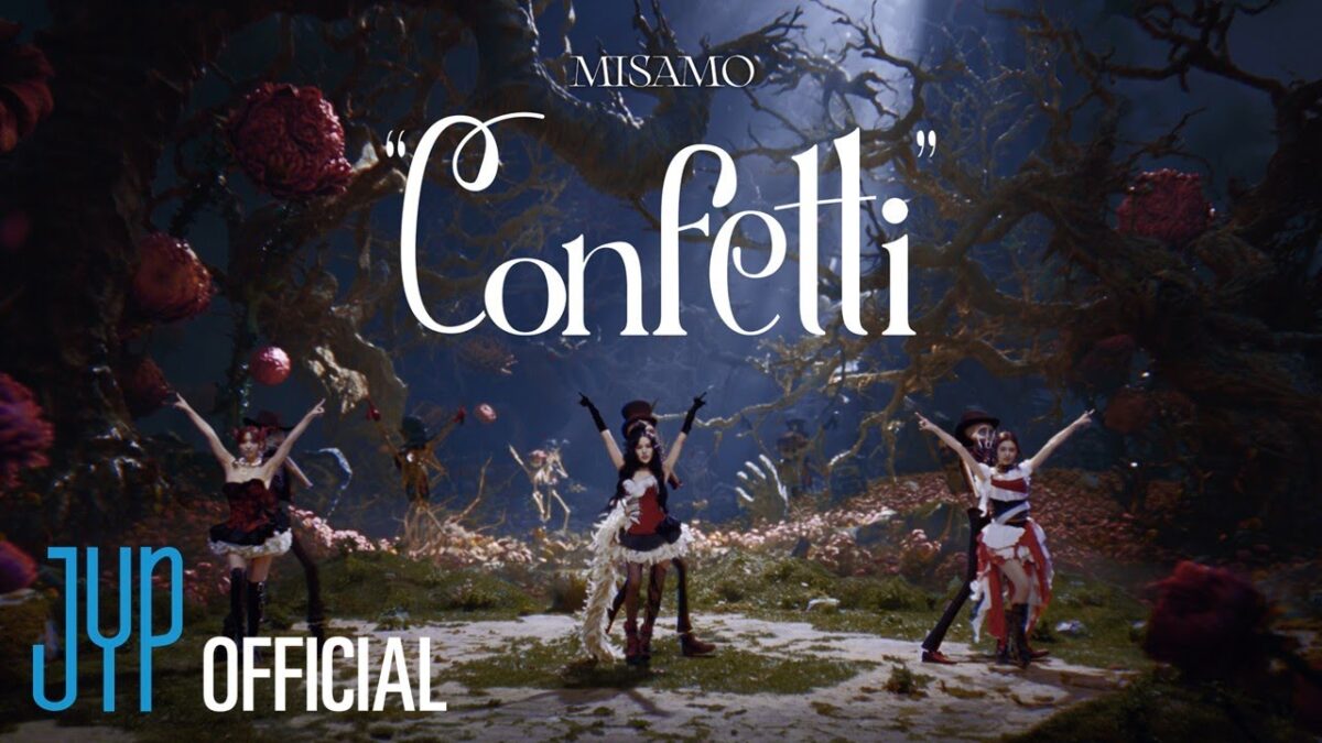 MISAMO se apresenta em um cenário mágico, dançando com figurinos elaborados e elementos de fantasia ao fundo.