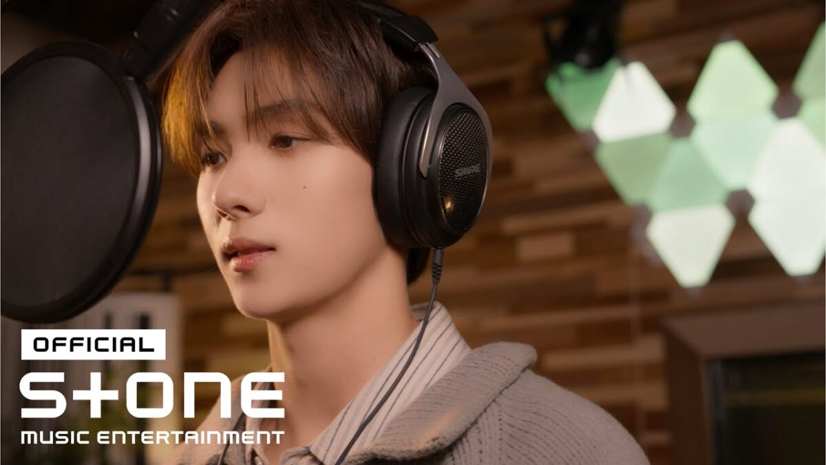 Youngjae grava em estúdio, usando fones de ouvido e se concentrando no microfone.