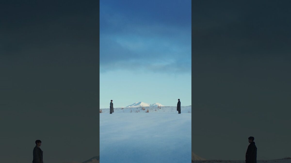 Dois homens em trajes escuros estão em um campo coberto de neve, com montanhas ao fundo sob um céu azul.
