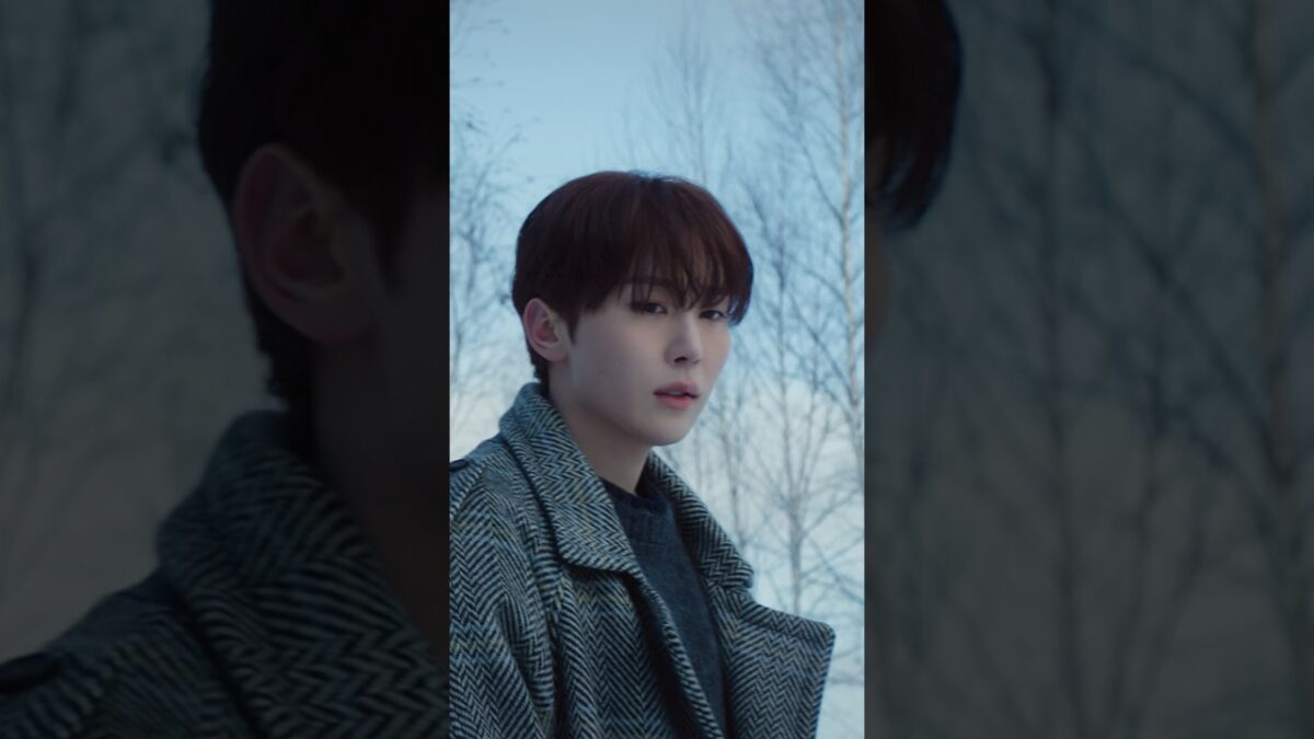 Dokyeom em um casaco estiloso, olhando para a câmera em um ambiente ao ar livre com árvores ao fundo.