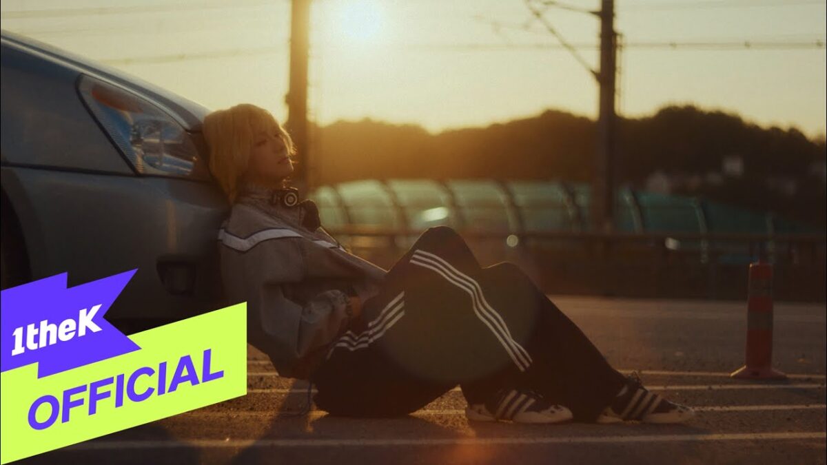 Uma jovem com fones de ouvido relaxa encostada em um carro enquanto o sol se põe ao fundo.