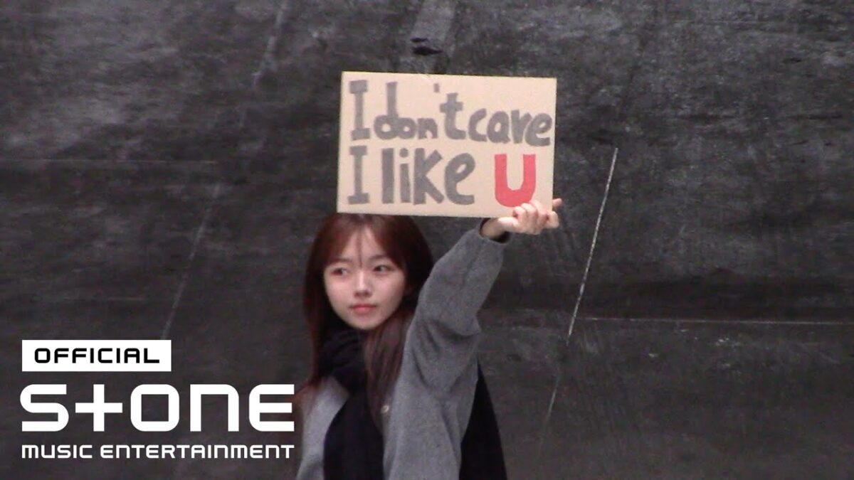 Jovem segura um cartaz escrito "I don't care I like U" em um cenário urbano.