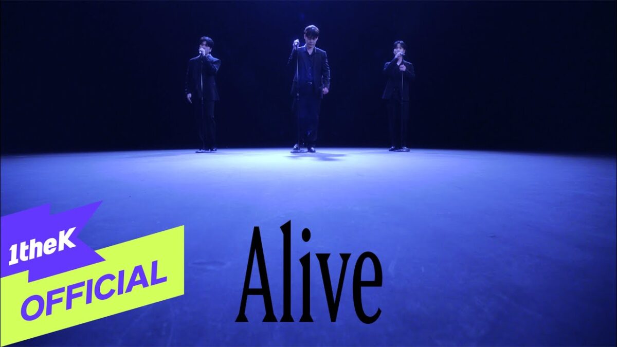Três cantores em trajes pretos se apresentam em um palco iluminado com luz azul, destacando a palavra "Alive".