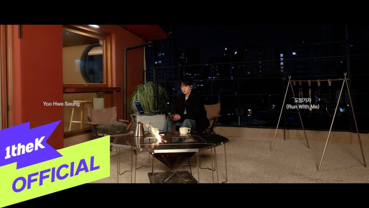 Yoo Hwe Seung sentado em uma varanda à noite, com uma mesa, fogo e decoração urbana ao fundo.