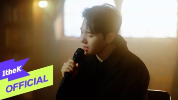 Kim Min Seok canta com emoção, segurando um microfone em um ambiente iluminado suavemente.