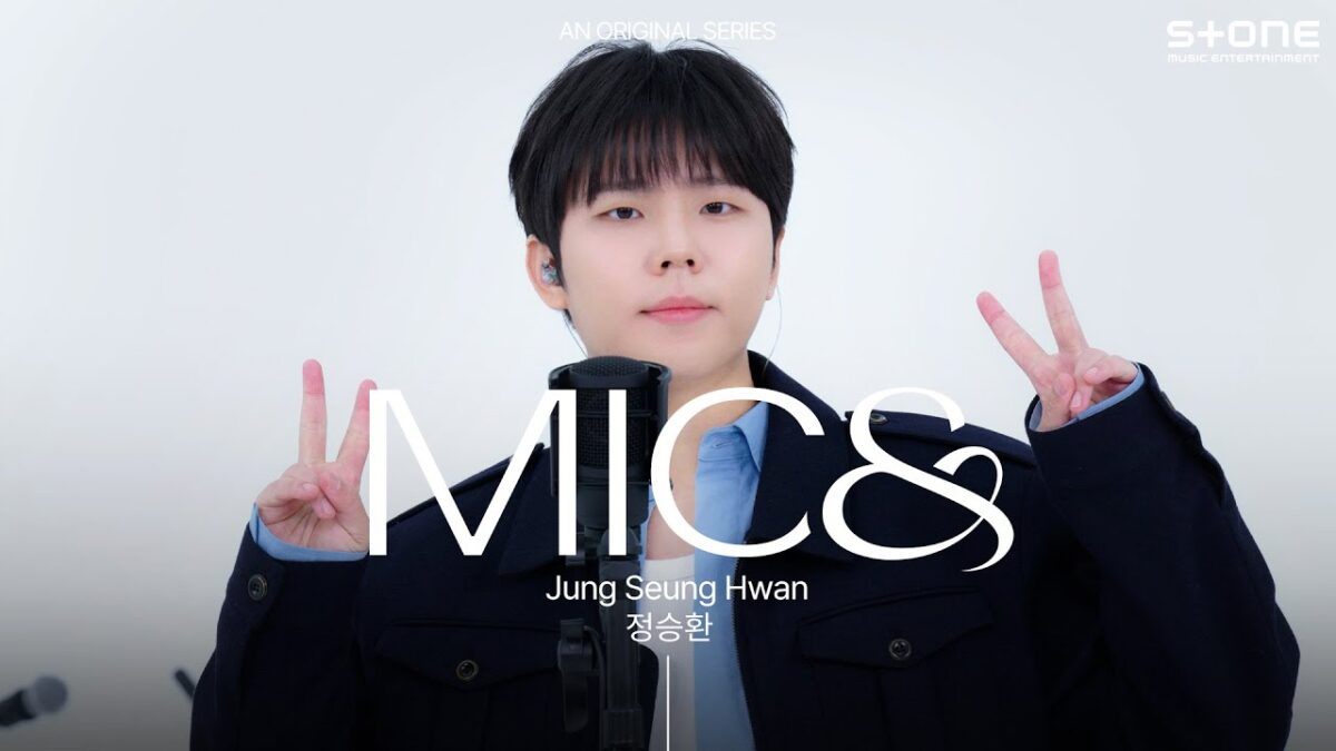 Jeong Seung Hwan faz sinal de paz, posando ao lado de um microfone em um ambiente claro e minimalista.