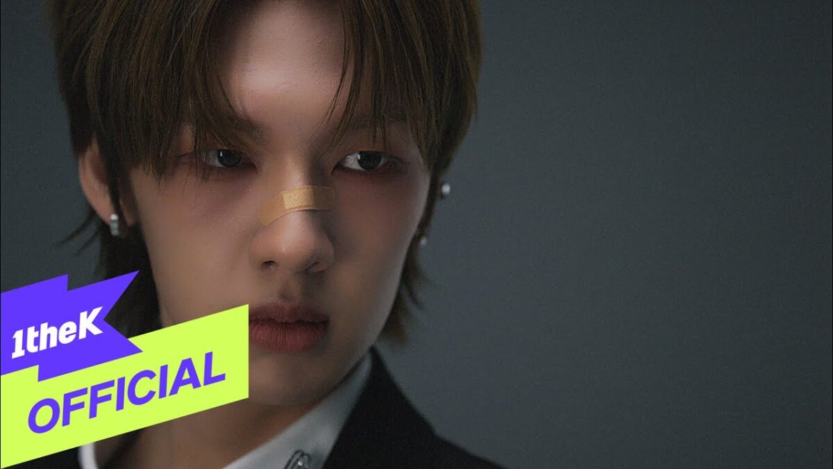 Jovem com curativo no nariz, olhar intenso, vestindo terno preto, em fundo neutro. Logotipo "1theK OFFICIAL" no canto.