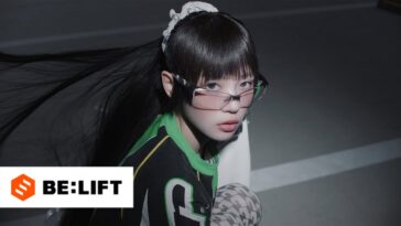 Artista com cabelo longo e liso, usando óculos e camiseta verde, olhando para a câmera em um ambiente escuro.