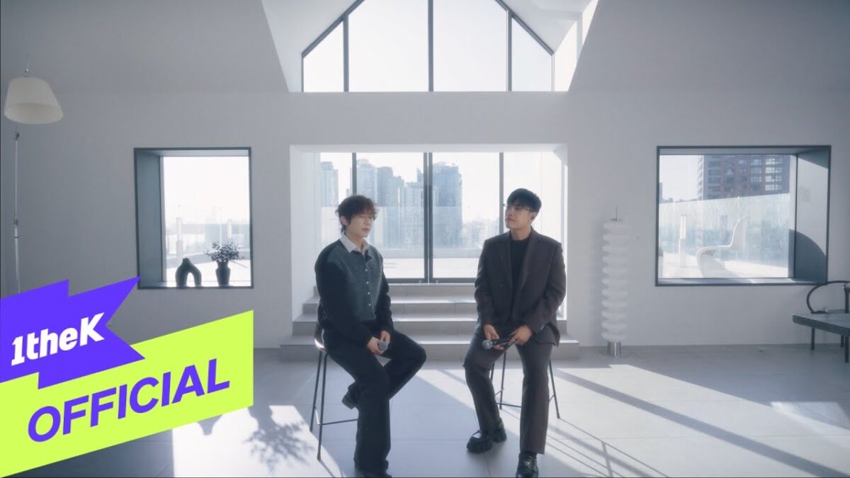 Song I Han e Jeon Sang Keun conversam em um ambiente moderno e iluminado, com vista para a cidade.