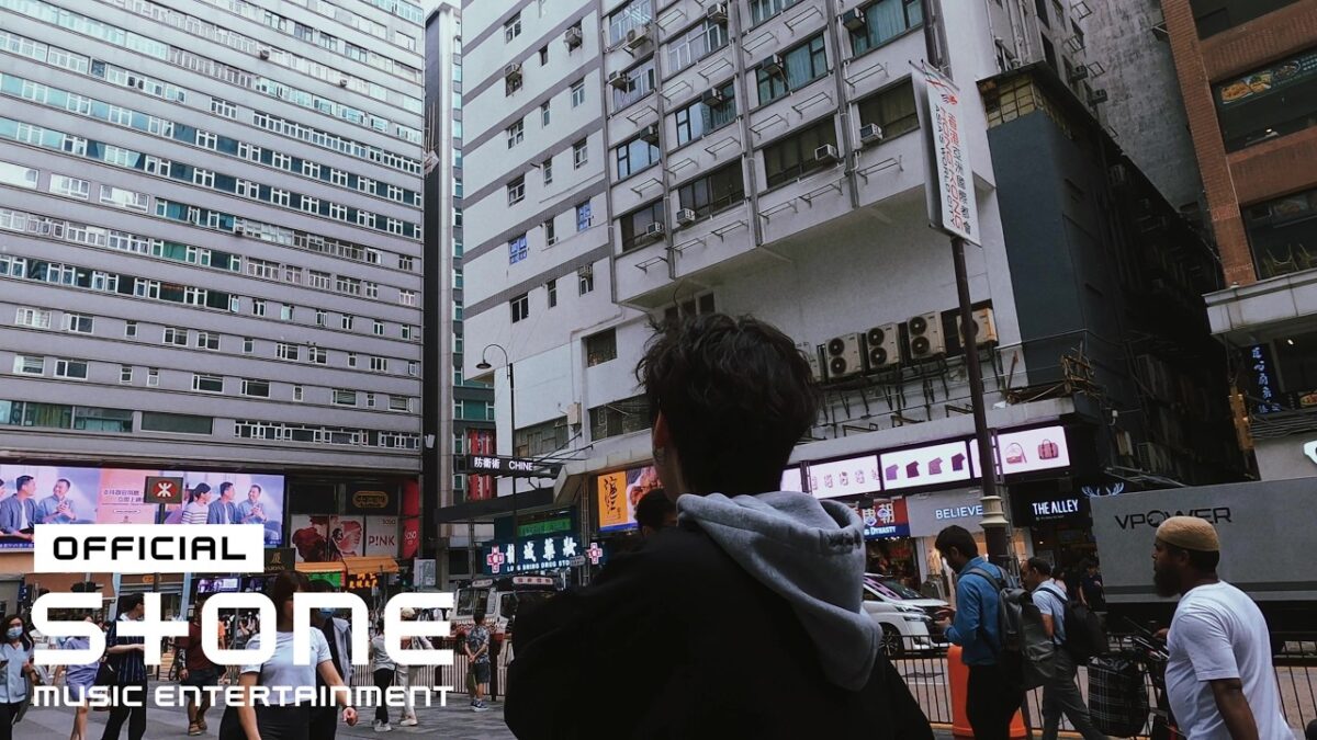 Jovem observa edifícios altos e movimentação de pessoas em uma rua de Hong Kong, com painéis publicitários visíveis.