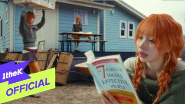 Uma jovem de cabelos laranjas lê um livro sentado em um pátio, enquanto outra pessoa carrega uma caixa ao fundo.