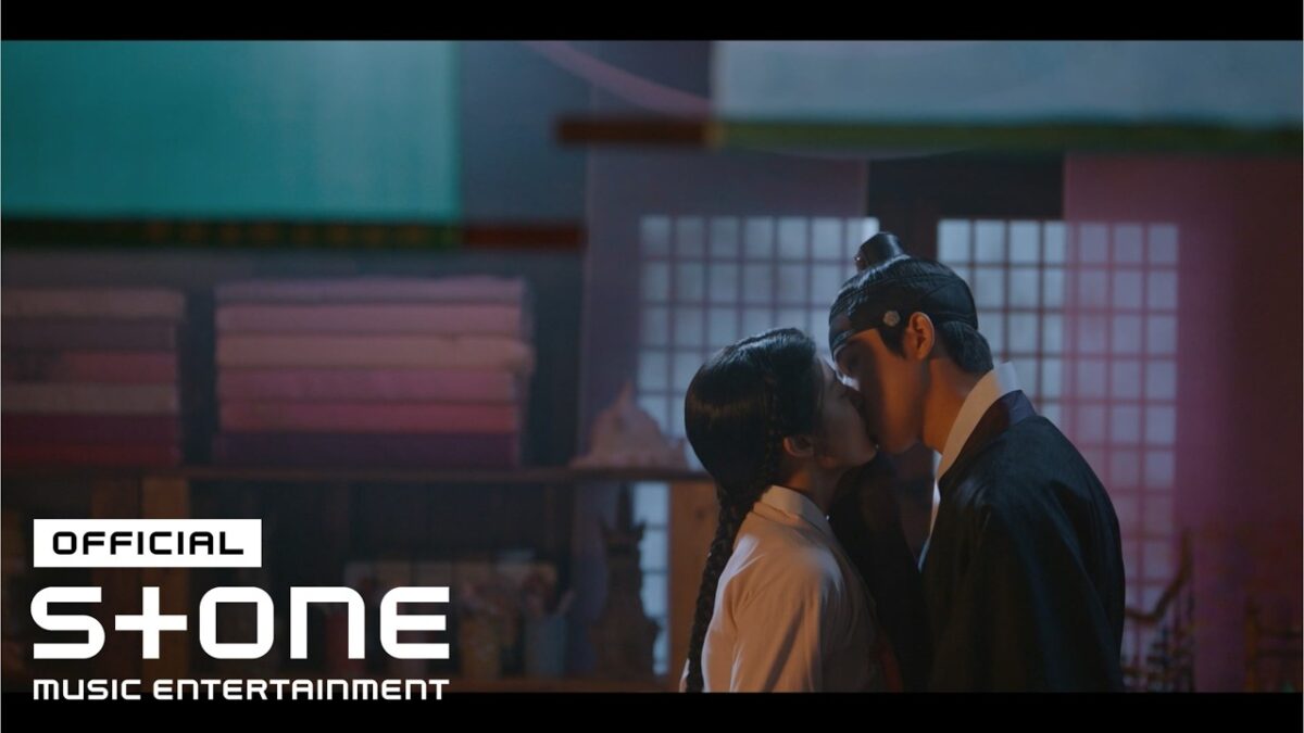 Casal se beijando em um ambiente romântico, com elementos decorativos ao fundo que remetem a uma época histórica.