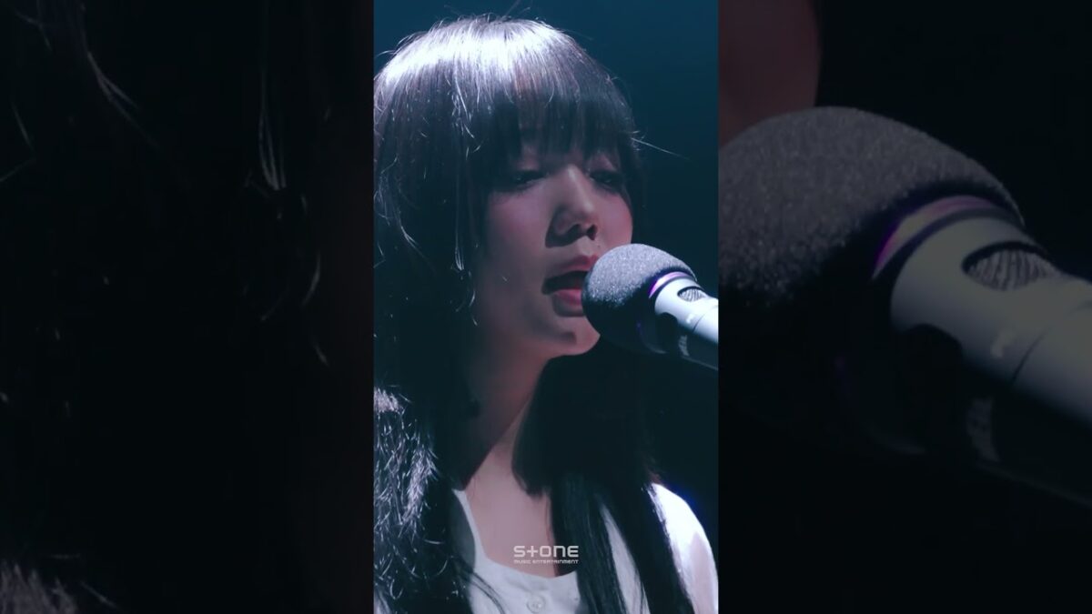 Cantora com cabelo longo e liso, cantando em um microfone durante apresentação ao vivo, com iluminação suave.