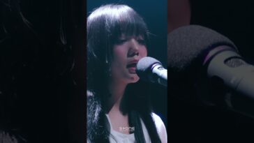 Cantora com cabelo longo e liso, cantando em um microfone durante apresentação ao vivo, com iluminação suave.