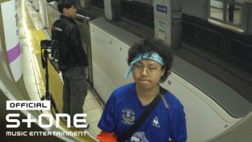 Dois jovens em uma estação de metrô, um deles com cabelo cacheado e bandana, o outro ao fundo com uma câmera.