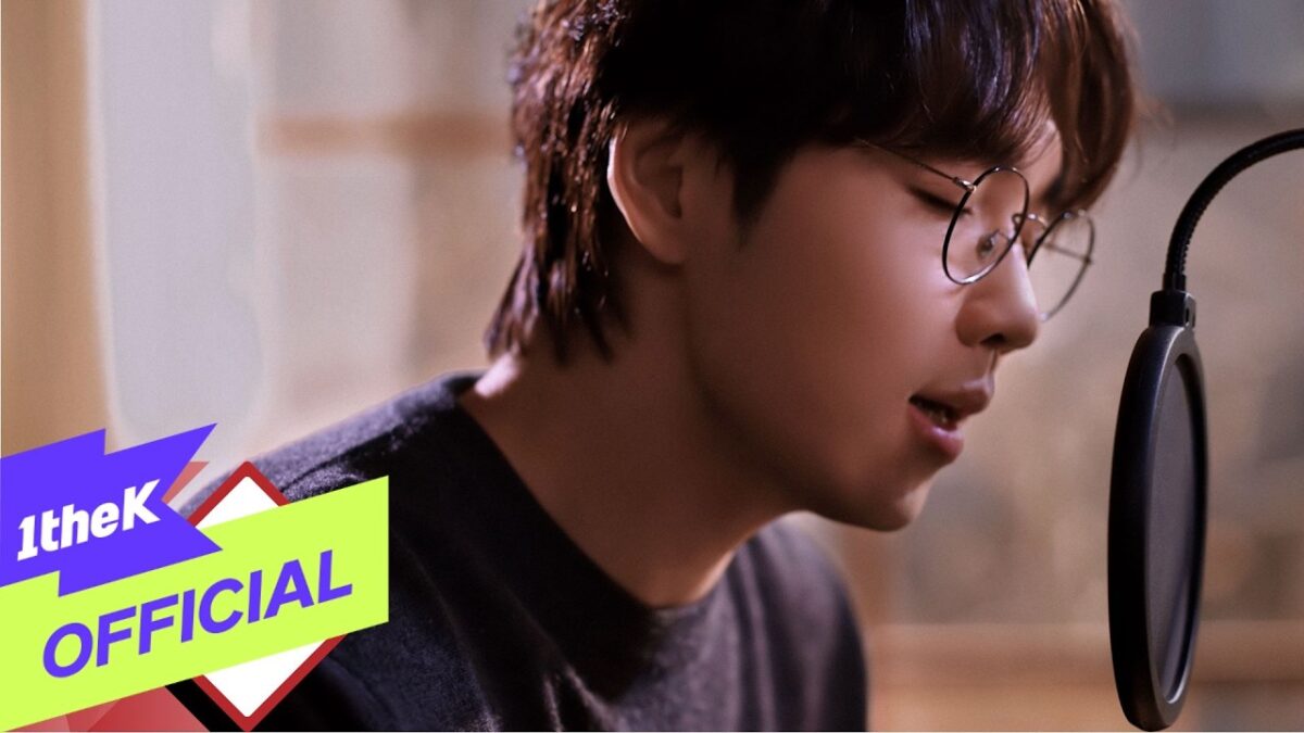 Lee Seok Hoon canta em um estúdio, usando óculos, com um microfone próximo ao rosto.