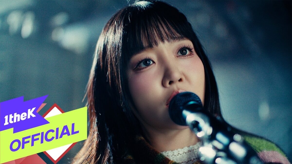 Younha canta emocionada em um palco, com um microfone próximo ao rosto e iluminação suave ao fundo.
