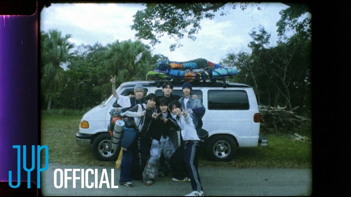 Sete jovens posam alegres em frente a uma van branca, com mochilas e equipamentos de camping no teto.