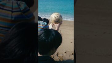 Uma pessoa segura uma bola de futebol com um sorriso desenhado, em frente a uma praia com água azul.