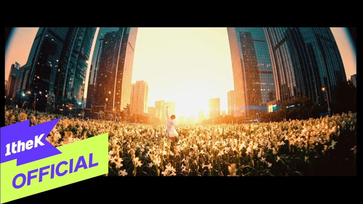 Homem vestido de branco dança em um campo florido, com arranha-céus ao fundo e o sol poente iluminando a cena.