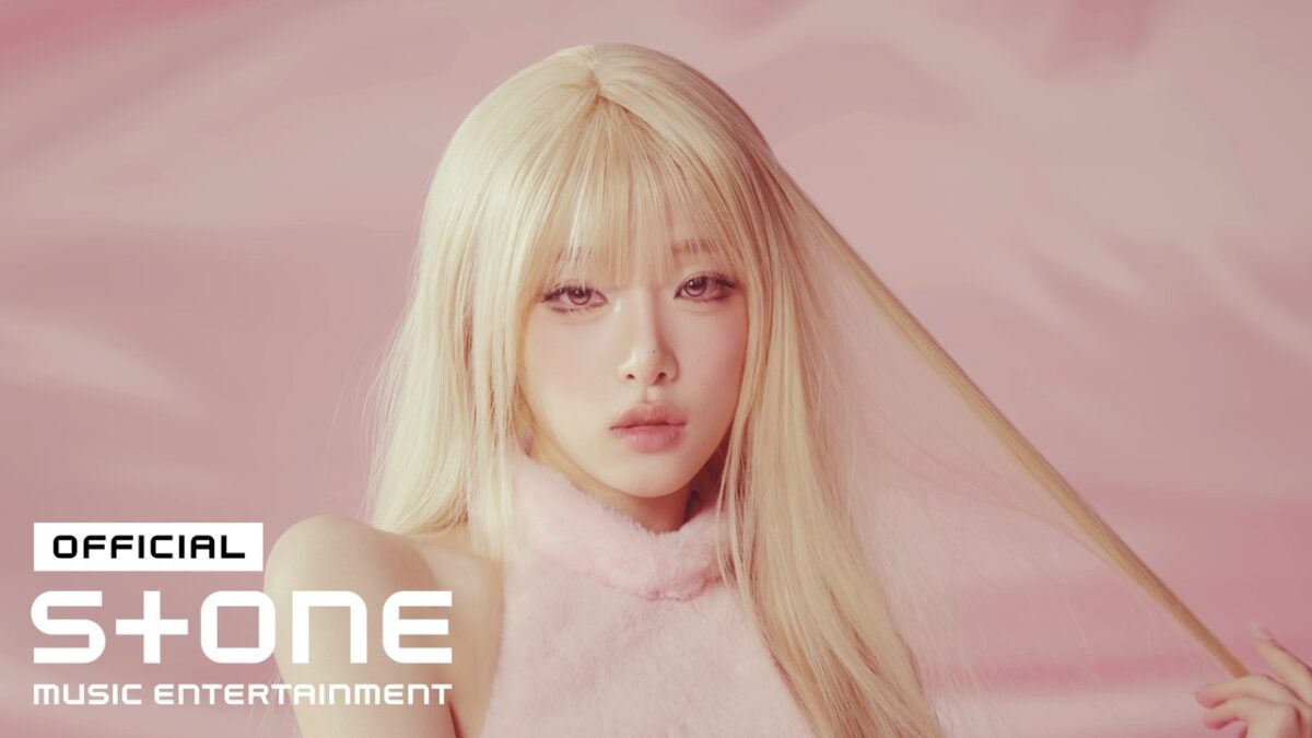 Artista com cabelo longo e liso, usando um top rosa, em fundo suave e rosa, olhando diretamente para a câmera.