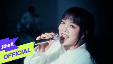 Younha canta empolgada com microfone em mãos, usando uma blusa branca e um detalhe de borboleta no rosto.