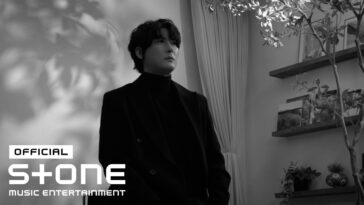 Jungsuk posa em preto e branco, vestido de preto, em um ambiente com plantas e prateleiras decorativas ao fundo.